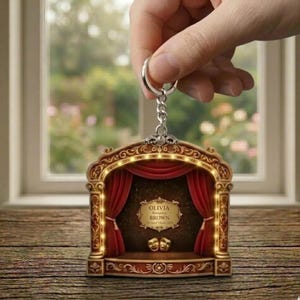 May include: A theatrical stage-shaped keychain with a red curtain, gold accents, and the text "OLIVIA BROWN". The keychain has a silver chain and ring. The stage has two theater masks and decorative lights.