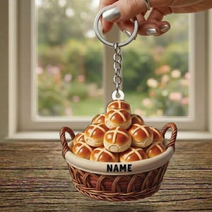 May include: A keychain featuring a miniature basket filled with miniature hot cross buns. The basket is brown with a white interior and the word "NAME" printed on the front. The buns are golden brown with a cross design.