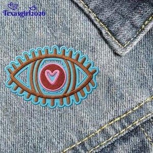 May include: An embroidered patch featuring an eye design with a brown iris, a pink center, and a light blue heart. The patch has a light blue scalloped border with brown radiating lines and is placed on blue denim fabric with the text texasgirl2026.