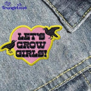 May include: Embroidered patch with a pink heart shape, yellow border, and the text "LET'S GROW GIRLS!" in black. Two black crow illustrations are on either side of the heart. The patch is set against a denim background.