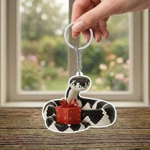 May include: A keychain featuring a stylized snake with a black and white pattern, holding a red gift box with a red bow. The keychain is attached to a silver ring and chain. The background is a blurred view of a window and wooden surface.