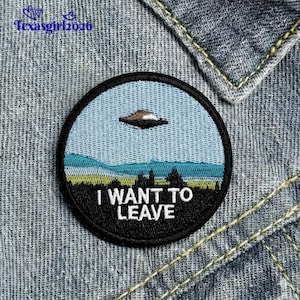 May include: Embroidered patch with a circular design featuring a landscape with a UFO and the text "I WANT TO LEAVE". The patch has a black border and is set against a light blue sky and green and blue landscape.
