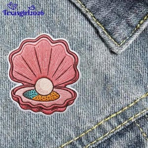 May include: An embroidered patch featuring a pink open seashell with a central white pearl and a small colorful patterned section at the base. The patch is applied to a blue denim fabric background with visible yellow stitching and the text texasgirl2026.