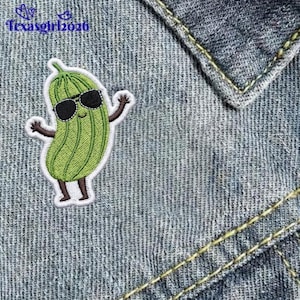 May include: An embroidered patch featuring a light green pickle character with a smiling face and black sunglasses. The pickle has brown arms and legs extended outward. The patch is outlined in white and is placed on blue denim fabric.