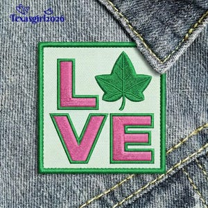 May include: Embroidered patch with the word "LOVE" in pink letters with green outlines, and a green ivy leaf. The patch is on a light green background, framed by a green border, and set against a denim background.