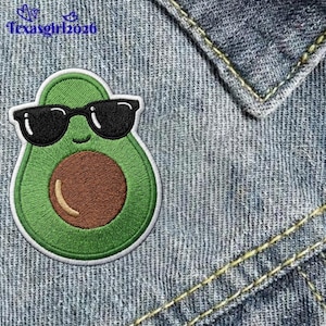 May include: An embroidered patch shaped like a green avocado wearing black sunglasses with a brown pit in the center. The patch features a white border and is placed on blue denim fabric. The text texasgirl2026 is visible in the top left corner.