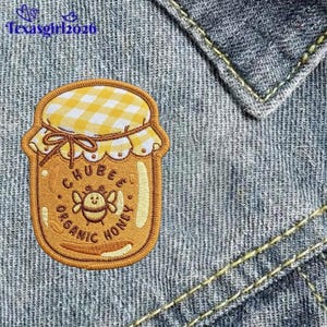 May include: An embroidered patch shaped like a honey jar with a yellow and white gingham lid. The jar is golden brown with a central bee graphic and the text CHUBEE ORGANIC HONEY. The patch is attached to a blue denim fabric with yellow stitching.