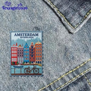 May include: A rectangular patch featuring an embroidered illustration of Amsterdam, Netherlands, with colorful canal houses, a bicycle, and a boat. The text reads AMSTERDAM NETHERLANDS. The patch is attached to a textured blue denim fabric.