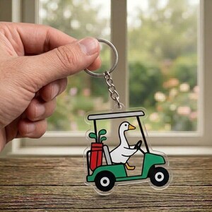 May include: A clear acrylic keychain featuring a cartoon goose driving a green golf cart. The golf cart has black wheels and a red golf bag with clubs. The goose is white with a yellow beak. The keychain is held by a silver keyring.