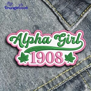 May include: Embroidered patch with the words "Alpha Girl 1908" in green and pink. The patch has a white background and is outlined in pink. Two green ivy leaves are on the bottom.