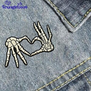 May include: An embroidered patch featuring a skeleton hand forming a heart shape. The patch has a black border with off-white bone details. The patch is on a denim background.