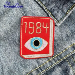 May include: A red embroidered patch featuring the year "1984" in gold thread, with an eye design below. The eye has a blue iris, a black pupil, and a pink outline. The patch is rectangular and set against a denim background.