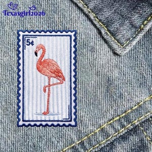 May include: A rectangular embroidered patch featuring a pink flamingo on a light blue and white striped background, designed to resemble a postage stamp with a 5 cent denomination, stitched onto a blue denim fabric with yellow stitching.