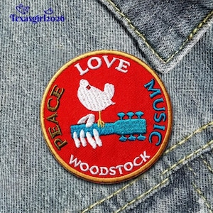 May include: A round, red patch with gold trim featuring a white dove perched on a blue guitar. The words "LOVE", "PEACE", "MUSIC", and "WOODSTOCK" are embroidered in white and blue. The patch is set against a denim background.