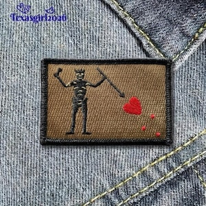 May include: A rectangular patch featuring a brown background with a black skeleton figure holding a spear and a red heart with blood drops. The patch has a black border and is set against a denim background.
