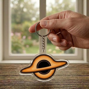 May include: A clear acrylic keychain featuring a black hole design with orange and brown concentric rings. The keychain is attached to a silver metal ring and chain. The design is held up by a hand.