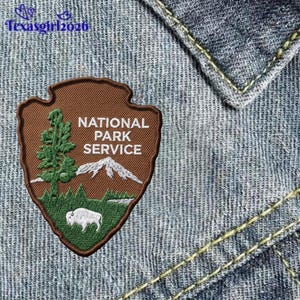 May include: A brown shield-shaped embroidered patch featuring the National Park Service logo with a green tree, white mountain peaks, and a white bison on a green field. The text reads National Park Service in white capital letters.
