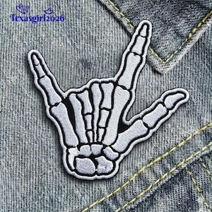 May include: Embroidered patch featuring a white skeleton hand in the 'rock on' gesture, outlined in black. The patch is set against a denim background, showcasing a detailed design with a stitched border.