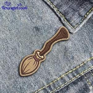 May include: Embroidered patch of a cartoon broom with a brown handle and tan bristles. The patch is outlined in a tan color and is set against a denim background. The text "Texasgirl2026" is in the upper left corner.
