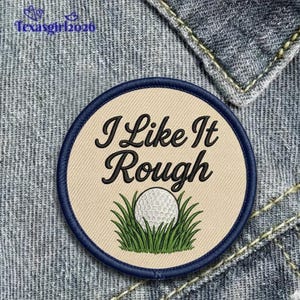 May include: Embroidered patch with the text "I Like It Rough" in black script. The patch features a golf ball in green grass against a tan background, framed by a navy blue border. The patch is on a denim background.