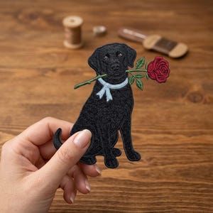 May include: An embroidered patch featuring a black Labrador retriever sitting down. The dog has a light blue ribbon tied around its neck and is holding a single red rose with a green stem and leaves in its mouth. The patch has detailed stitching.