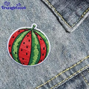 May include: A round embroidered patch featuring a stylized watermelon design with alternating red and green vertical segments and small black seed details, attached to a blue denim fabric background with the text texasgirl2026 visible.