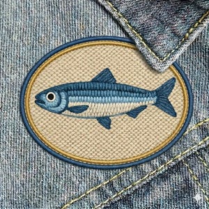 May include: Embroidered patch featuring a blue fish design. The fish is detailed with light blue and white stripes, set against a beige background within a navy blue and gold oval. The patch is sewn onto a denim fabric.