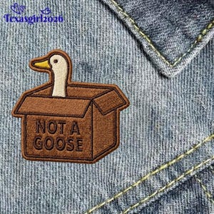 May include: An embroidered patch featuring a white goose head with a yellow beak emerging from a brown cardboard box. The box has the text NOT A GOOSE embroidered in dark brown thread on the front. The patch is attached to blue denim fabric.