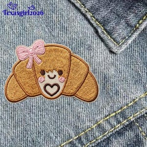 May include: An embroidered patch featuring a tan croissant with a smiling face, pink cheeks, and a brown heart on its center. A light pink bow is attached to the top left side of the croissant. The patch is placed on blue denim fabric.