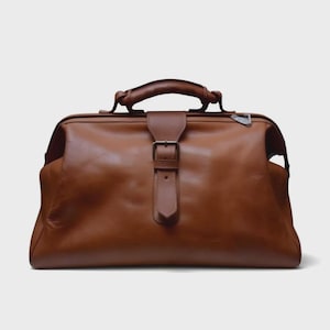 May include: A brown leather doctor's bag with a top handle and a buckle closure. The bag has a rectangular shape and a classic design. The leather appears to be smooth and has a rich, warm color. The bag is suitable for carrying various items.
