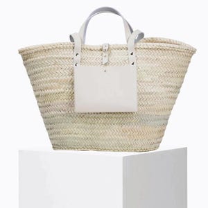 May include: A woven straw tote bag with white leather handles and a white leather pocket. The bag has a rectangular shape with a wide opening and a light-coloured woven pattern. The handles are attached with silver rivets.