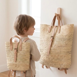 May include: A natural woven straw backpack with tan leather straps and a buckle closure. The backpack has a rectangular shape and a top handle. A larger matching tote bag hangs on the wall. A young child is wearing the backpack.