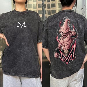 May include: Dark gray t-shirt with a distressed finish. The front features a white "M" logo. The back displays a colorful graphic of a character with pink skin, pointing, and a pink and red "M" logo. The shirt has short sleeves.