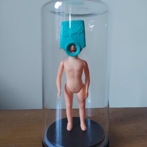 Peut inclure: Une poupée en plastique de couleur chair avec un couvre-chef turquoise à l'intérieur d'une cloche en verre transparent. La poupée se tient sur une base circulaire noire. La cloche est sur une surface en bois.