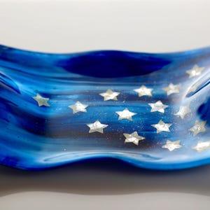 May include: A rectangular glass dish with wavy, upturned edges featuring a deep blue and light blue marbled pattern. The surface is decorated with multiple small, silver-toned, five-pointed star shapes scattered across the center of the dish.