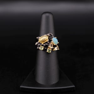 May include: A black and gold ring with a cluster of green, blue, and white gemstones. The ring is set on a black ring stand.