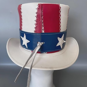 Puede incluir: Un sombrero de copa hecho de paneles de cuero rojo, blanco y azul. La corona cilíndrica presenta rayas verticales rojas y blancas con costuras blancas, mientras que la banda azul muestra estrellas de cuero blanco y un cordón de cuero.