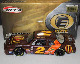 May include: A brown and orange die-cast model car with the number 2 on the roof and sides. The car has sponsor logos and Goodyear tires. The box behind the car has the RCCA logo and the word "ELITE".