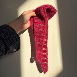Puede incluir: Bufanda tejida a mano en tonos rojos y burdeos. La bufanda tiene un diseño de tejido abierto y texturizado. La lana suave y esponjosa crea un accesorio acogedor y cálido. La bufanda se sostiene contra una pared blanca, con sombras.