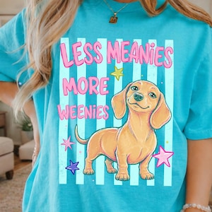 May include: Turquoise t-shirt with the text "LESS MEANIES MORE WEENIES" in pink. A cartoon dachshund dog is centered below the text, with stars in pink, yellow, and purple. The shirt has a relaxed fit.