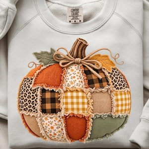 Puede incluir: Una sudadera de cuello redondo gris claro con un gran gráfico central de calabaza hecho de varios parches de tela. El diseño de la calabaza incluye cuadrados naranjas, amarillos, marrones y verdes con estampados florales, de cuadros y leopardo.