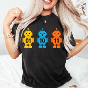 May include: Black t-shirt featuring three colorful robot graphics. The robots are yellow, blue, and orange, each with a unique expression and number: 01, 10, and 11. The shirt is made of a soft material.