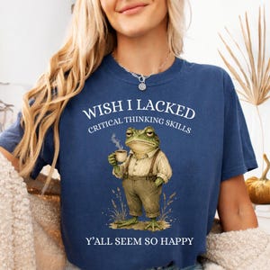 May include: A navy blue t-shirt featuring a graphic of a frog wearing a button-down shirt and suspenders while holding a coffee cup. The text reads WISH I LACKED CRITICAL THINKING SKILLS Y'ALL SEEM SO HAPPY in white lettering above and below the frog.
