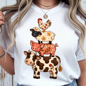 May include: White t-shirt featuring a whimsical illustration of a cow, pig, sheep, and chicken stacked on top of each other. The cow is brown and white, the pig is pink, the sheep is white, and the chicken is beige with red accents. The design has a floral pattern.