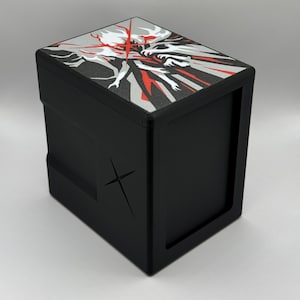 May include: Black box with a graphic design on the top featuring red, white, and silver elements. The box has a cross-shaped design on the side. The box is likely for storing small items.