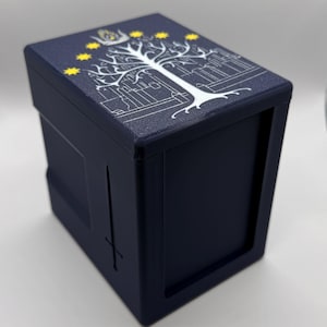Può includere: Una scatola blu navy con un design dettagliato di albero bianco e stelle sul coperchio. La scatola ha un design a spada sul lato. Il design ricorda un tema fantasy.