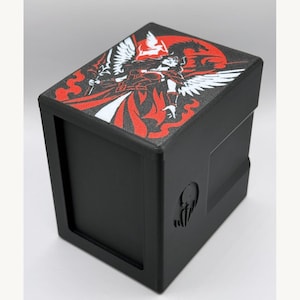 May include: A black rectangular deck box featuring a top lid with a graphic illustration of a winged figure in red, white, and black. The side of the box displays a small embossed circular emblem. The structure is solid black with a matte finish.