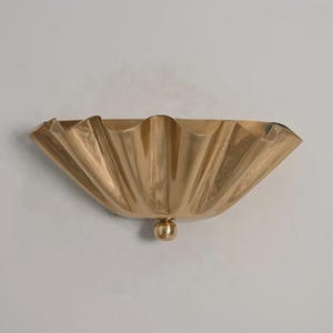 May include: A gold-colored, wall-mounted decorative bowl with a scalloped rim and a small, round base. The bowl has a smooth, reflective surface and is mounted on a white wall.