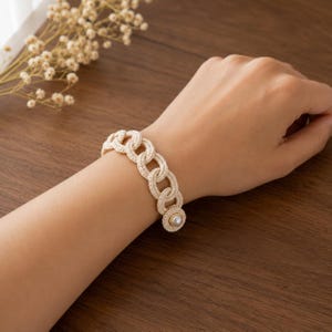 Può includere: Un braccialetto all'uncinetto color crema caratterizzato da una serie di maglie circolari interconnesse. Il braccialetto è fissato con un unico bottone rotondo dall'aspetto perlato all'estremità, creando un design a maglie strutturato.