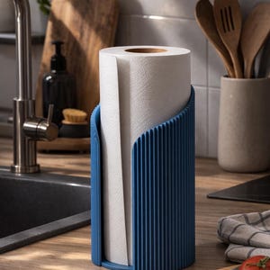 May include: A cylindrical paper towel holder featuring a deep blue color and a vertical ribbed texture. The holder is open on one side to allow for the insertion of a standard roll of white paper towels, which is currently positioned inside the unit.